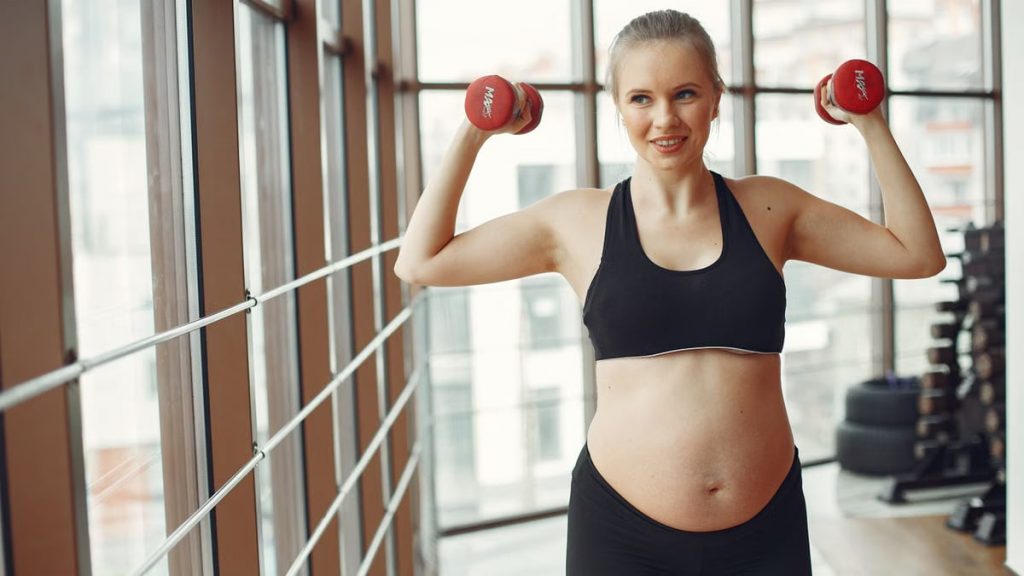 Expectant woman in workout gear lifts dumbbells indoors, embodying fitness and health.